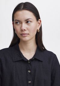 Camicia nera con bottoni, colletto, tessuto texturizzato e orecchini ad anello dorati. Il modello ha i capelli lunghi e lisci e un'espressione neutra.