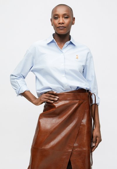 Pasitikintis žmogus, vilkintis šviesiai mėlyną marškinių palaidinę su mažuoju lapės siuvinėjimu ir rudą odinę sijoną, ranka ant klubo prieš baltą foną.