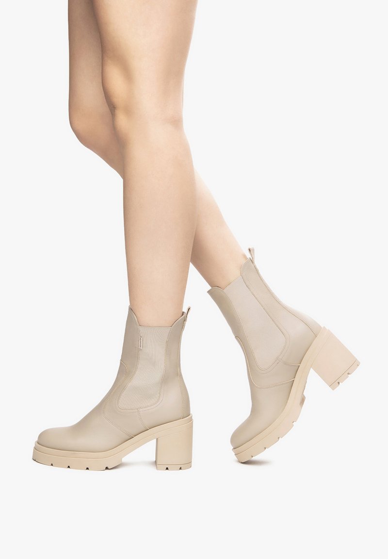 Beige Knöchelstiefel mit einem dicken Absatz, der über elastische Seitenpaneele und eine glatte Textur verfügt. Robuste Sohle mit Profilierung für besseren Halt.