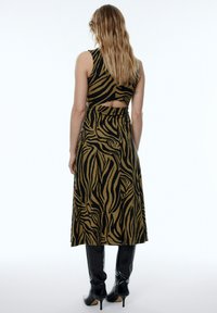 Maxi-Kleid in einem braun-schwarzen Zebra-Muster, mit einem ausgeschnittenen Rückendesign, tailliertem Oberteil und fließendem Rock. Getragen mit kniehohen schwarzen Stiefeln.