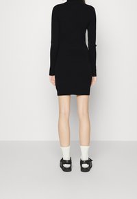 Robe noire à manches longues côtelées, silhouette ajustée, longueur mi-cuisse. Portée avec des chaussettes blanches montantes et des sandales noires avec des détails à bride.