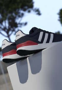 Par de zapatillas Adidas negras, blancas y rojas exhibidas sobre una superficie blanca con árboles y cielo desenfocados al fondo.