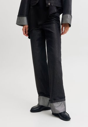 Le modèle porte un jean noir à jambes larges avec des revers gris oversize, un haut noir et des mocassins noirs.