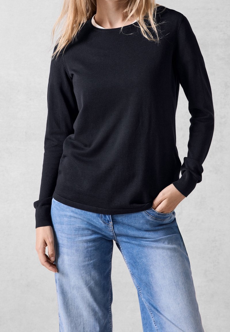 Personne portant une chemise noire à manches longues et un jean bleu clair, debout avec la main gauche dans la poche et la main droite détendue le long du corps.