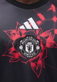 Το έμβλημα της ομάδας ποδοσφαίρου Manchester United και το λευκό λογότυπο της Adidas σε αθλητική φανέλα με μαύρο και κόκκινο φλοράλ μοτίβο.