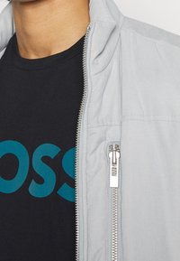 Ljusgrå zip-up jacka med hög krage, med silverdetaljer och en svart T-shirt under med turkosa bokstäver.