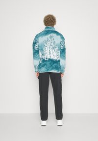 Veste en polaire turquoise avec imprimé forêt blanche au dos ; design à col et coupe décontractée. Associée à un pantalon noir et des baskets blanches.