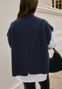 Frau in einem dunkelblauen, ärmellosen Strickwesten über einem weißen Hemd, die eine schwarze geflochtene Handtasche hält und im Freien neben einem weißen Gebäude steht.