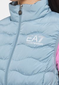 Smanicato imbottito azzurro chiaro con collo alto, design trapuntato orizzontale, chiusura con zip e logo EA7 Emporio Armani bianco. Rifinito con dettagli rosa.