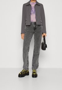 Giacca grigia cropped su un top trasparente viola, abbinata a jeans grigi dritti e stivali platform chunky neri e metallici dorati. Borsa nera.