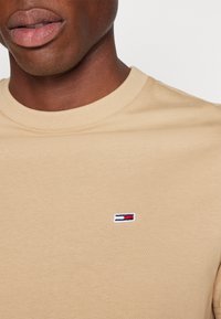 Béžové bavlněné tričko s kulatým výstřihem a krátkými rukávy, na hrudi s malým vyšitým logem v modré, bílé a červené barvě.