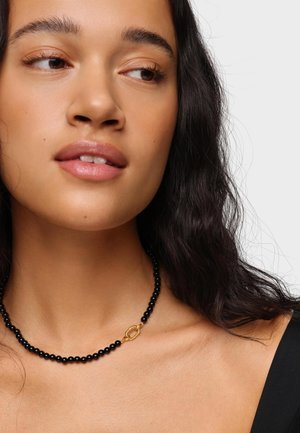 Joven mujer con cabello largo y oscuro que lleva un collar negro de cuentas con un cierre dorado, mirando ligeramente de lado sobre un fondo liso.