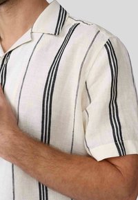 Chemise en lin à manches courtes couleur écrue avec des rayures verticales noires. Présente un col classique et un tissu texturé pour la respirabilité.