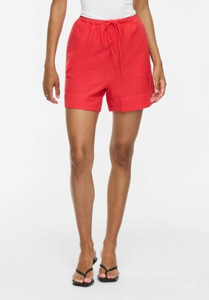 Kvinde iført lyse røde shorts med snøre og sorte sandaler med hæl, stående mod en hvid baggrund.