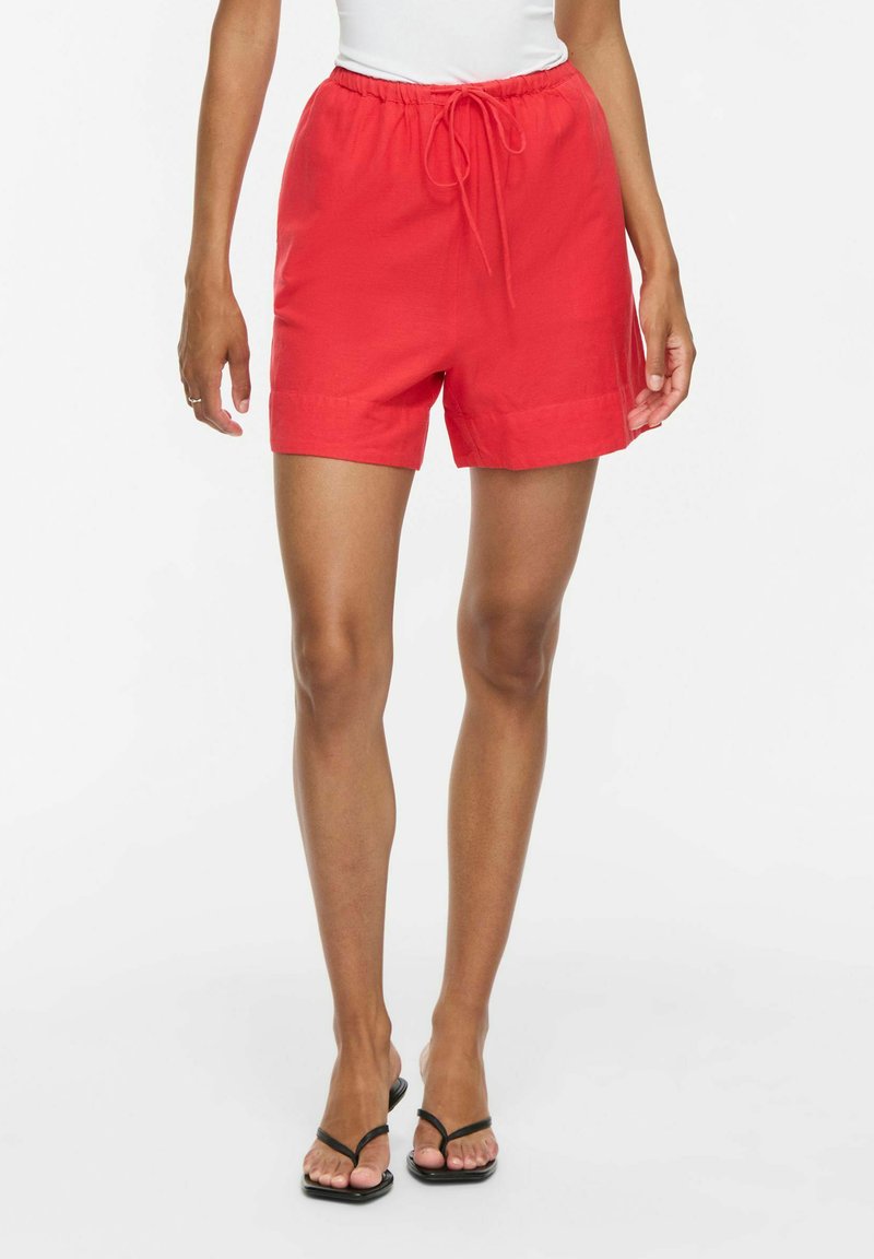 Femme portant un short rouge vif à cordon et des sandales à talons noires, debout devant un fond blanc.