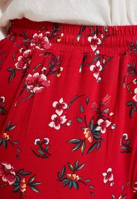 Červené kalhoty s elastickým pasem s květinovým vzorem v růžové, bílé, žluté a zelené barvě, nošené s bílým topem.