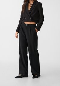 Vrouw in een zwart cropped blazer, wijde zwarte broek en puntige zwarte schoenen met studs straps tegen een effen achtergrond.