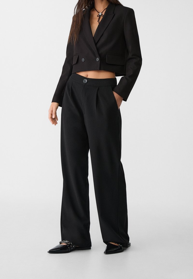 Vrouw in een zwart cropped blazer, wijde zwarte broek en puntige zwarte schoenen met studs straps tegen een effen achtergrond.