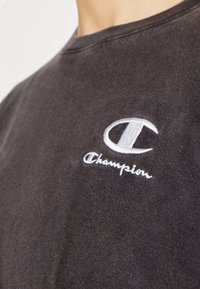 Tamno siva pamučna majica s bijelim izvezenim Champion logom na grudima. Klasični okrugli izrez i mekana tekstura.