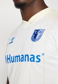 Hvid sportsjersey med lyse vertikale striber, prydet med blå branding "Humana" og "1.FC Magdeburg" logo, fremstillet af åndbart stof.