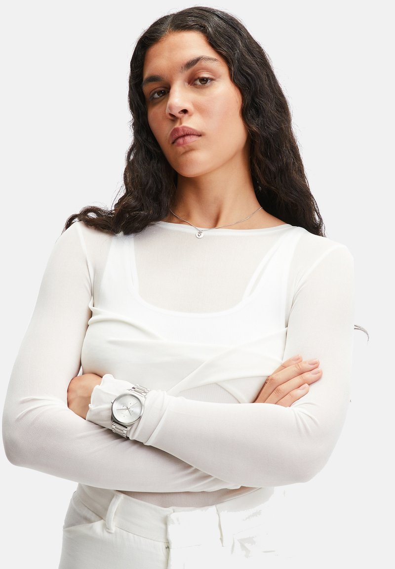 Weißes Langarmoberteil mit quadratischer Ausschnitt, kombiniert mit einer silbernen Uhr. Selbstbewusste Pose, Arme verschränkt, mit leicht welligem Haar.
