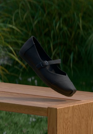 Schwarzer Mary Jane Schuh mit Schnallriemen, der über einer Holzbank schwebt, mit grünem Gras und Pflanzen im Hintergrund.