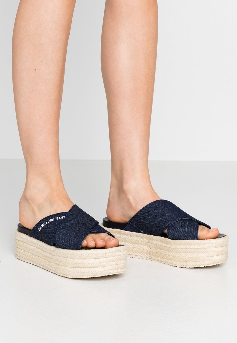 Pieds portant des sandales compensées bleu marine à brides croisées avec des semelles épaisses tissées beige sur un fond gris uni.