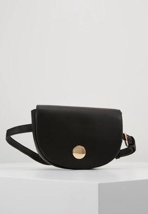Bolso de cinturón de cuero negro con solapa curva y cierre redondo dorado, colocado sobre una superficie blanca contra un fondo liso gris claro.