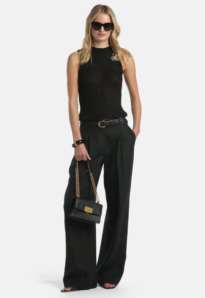 Plavolaska ženska, oblečena v črno brezrokavnik iz pletenine, široke črne hlače, črna sončna očala, v roki drži črno torbico z zlatim verižnim trakom.