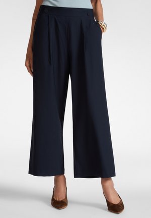 Femme portant un pantalon large court bleu marine, un haut bleu clair, des talons pointus marron et des bracelets au poignet droit.