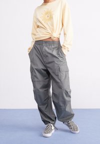 Person, der ein beiges Langarmhemd trägt, das um die Taille geknotet ist, graue Cargo-Hosen mit Taschen und schwarz-weiße kariertete Slipper auf einem blauen Boden.