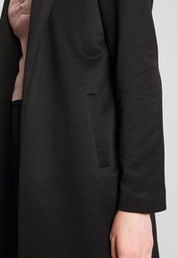 Manteau noir texturé avec un devant légèrement ouvert, doté d'une seule poche latérale cachée et d'un design de manches lisses.