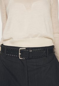 Schwarz geflochtener Leder Gürtel mit einer silbernen Schnalle, getragen über einem hellbeigen Oberteil. Das Design umfasst gleichmäßig verteilte Löcher und nähtechnische Akzente.