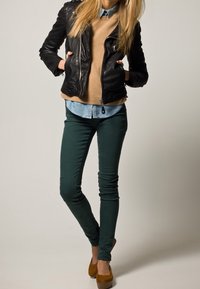 Veste en cuir noire, pull beige, chemise bleu clair, jean skinny vert, talons marron ; tenue décontractée avec des textures et des couleurs superposées.
