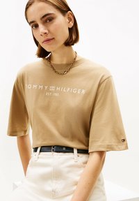 Beiges übergroßes T-Shirt mit weißem "TOMMY HILFIGER EST. 1985"-Logo. Kombiniert mit hellen Jeans und einem dunklen Gürtel, der einen Logo-Akzent setzt.