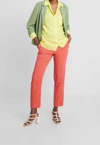 Veste verte, chemise boutonnée jaune, pantalon corail et talons blancs à brides. Textures lisses et designs ajustés avec des couleurs claires et lumineuses.