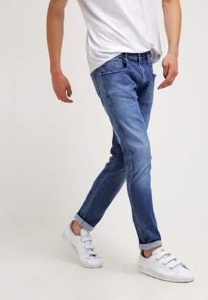 Ein Mann trägt aufgekrempelte blaue Jeans, weiße Sneakers mit Klettverschlüssen und ein weißes T-Shirt und geht vor einem einfarbigen hellen Hintergrund.