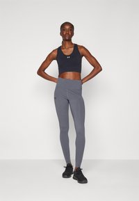 Mørkeblå sport BH og grå leggings med høy livlinje, tettsittende design og Under Armour-logo. Modellen har på seg svarte treningssko.