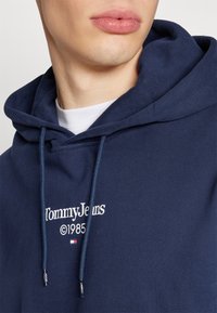 Námořnická modrá mikina z bavlněné směsi s kapucí na šňůrku a vyšitým logem "Tommy Jeans" s "©1985" a barevnými akcenty.