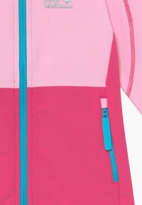 Fleecejacka i rosa och magenta, med en blå dragkedja och ficka. Den övre delen är ljusrosa, med kontrasterande sömmar.