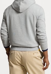Sudadera gris claro con capucha ajustable, puños y dobladillo acanalados. Llevada sobre pantalones beige con un bolsillo trasero con cremallera.