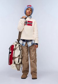 Kremowy sweter dziany z czerwono-czarnym wzorem flagi Norwegii, niebieska dziana czapka, koszula w kratę, beżowe spodnie typu cargo oraz duża wielokolorowa torba.