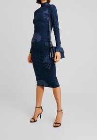 Navy, long-sleeve dress with floral pattern. Fitted midi design. Worn with black strappy heels and a small handbag. Smooth texture.