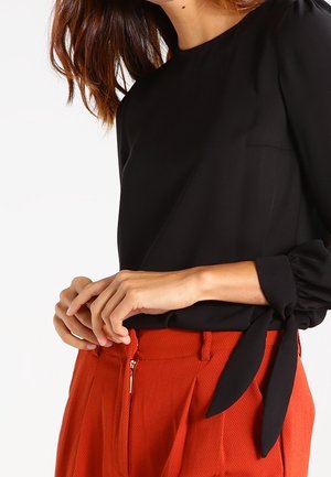 Vrouw draagt een zwarte top met lange mouwen met gestrikte manchetten en hooggetailleerde oranje textuurbroek met een rits aan de voorkant.