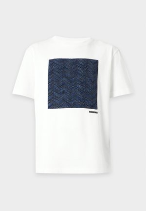 Junger Mann steht mit den Händen in den Taschen, trägt ein weißes T-Shirt mit blauem Quadrat-Print, schwarze Shorts und schwarze Slip-On-Schuhe.