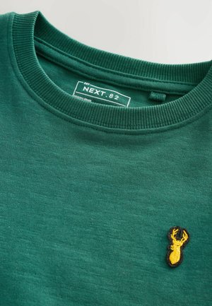 Sweatshirt vert avec un col rond, ornée d'un petit logo de cerf brodé jaune sur le devant. Tissu doux et texturé, style décontracté.
