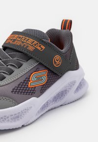 Zapatilla deportiva gris con acentos naranjas, material de malla y sintético, correa de Velcro y suela texturizada. Cuenta con función de luces en la suela.