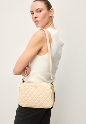 Naine, kes kannab valget varrukateta topi ja musti pükse, kannab õlal beeži tikandiga ristihoidjat ja vaatab üle õla.