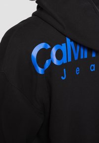Sudadera con capucha negra con un logo de texto azul reflectante grande y audaz que dice "Calvin Jeans" en el frente, mostrando una textura de tela suave.