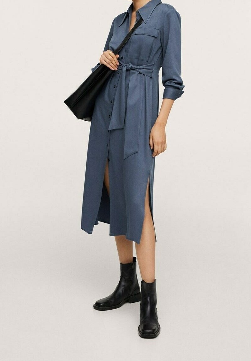 Abito camicia blu con maniche lunghe, fessure laterali e cintura, abbinato a stivali neri alla caviglia e a una borsa nera a tracolla.